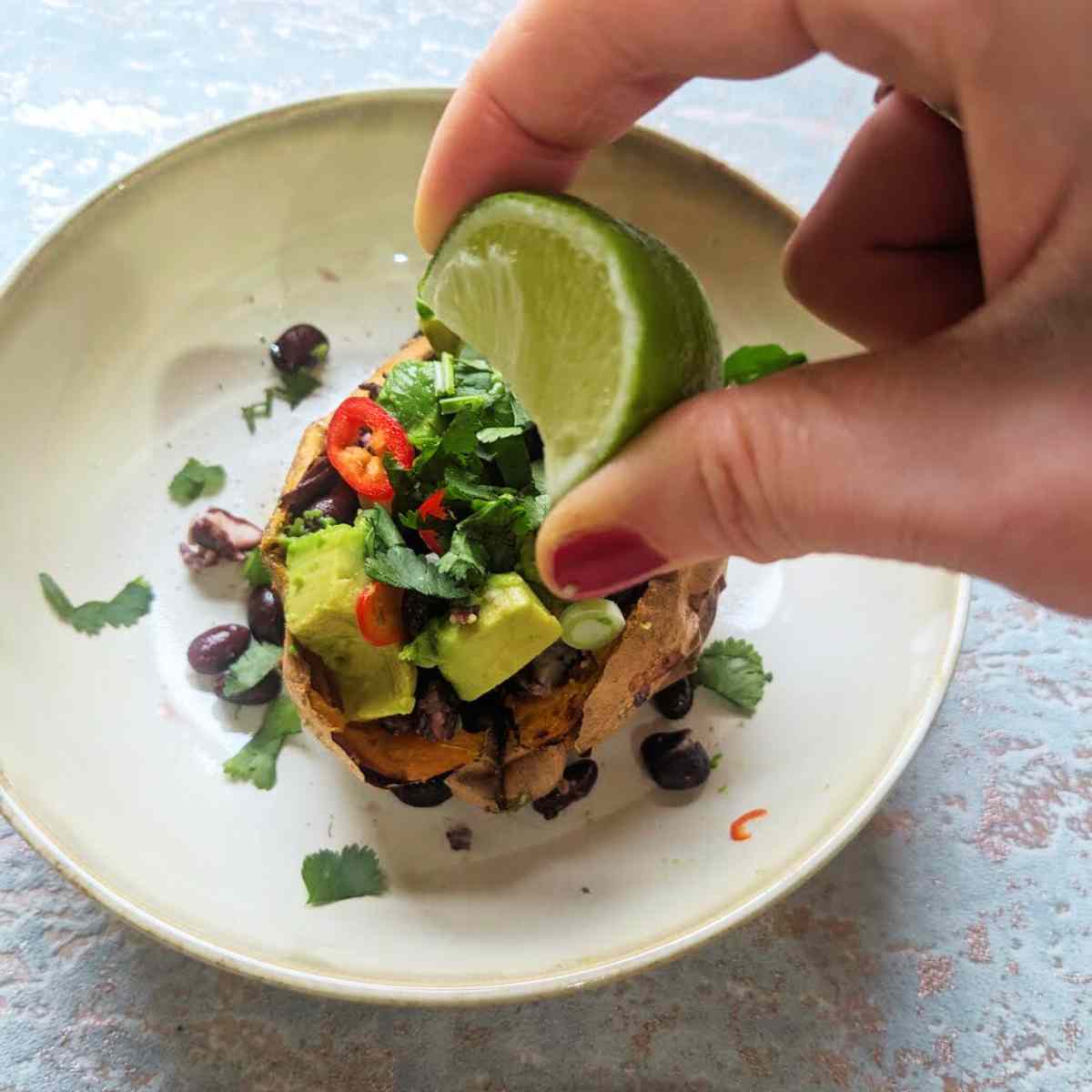 A hand is squeezing a wedge of fresh lime onto a baked sweet potato.