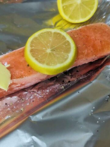 A frozen salmon fillet on a piece of aluminium foil with a slices of lemon and a pat of butter on top.