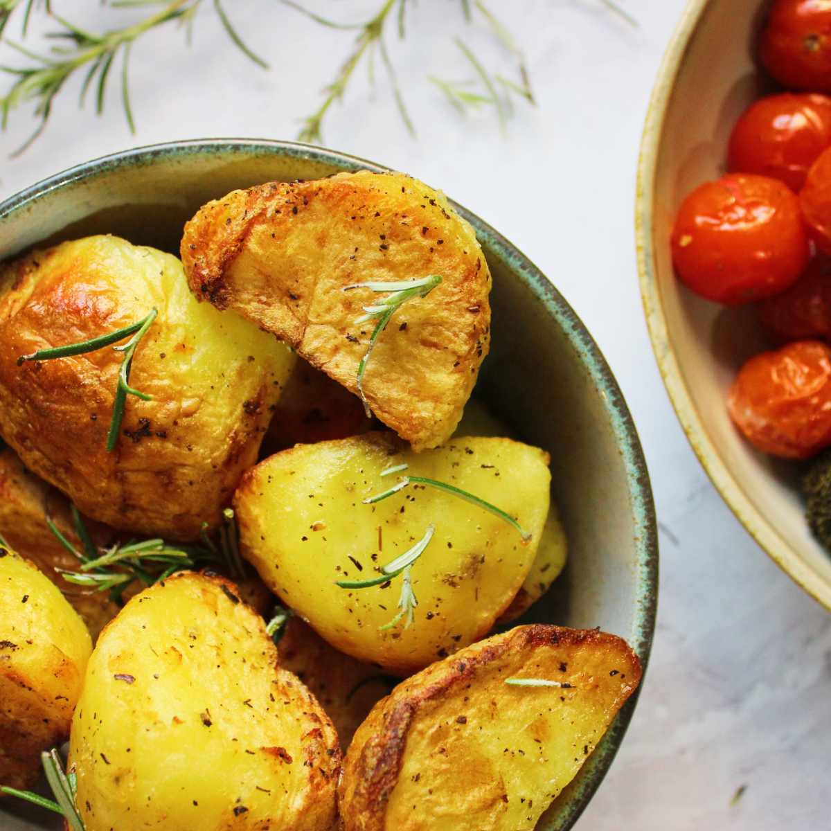 Roast potatoes topped with sprigs of rosemary in a bowl. A bowl of tomatoes sits on the side.