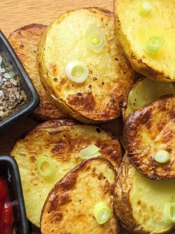 Sliced potatoes on a board topped with sliced scallions. Seasoning and tomato ketchup is in small bowls alongside.