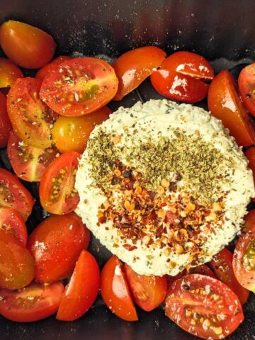 A round of Boursin cheese in an air fryer basket surrounded by halved plum tomatoes.