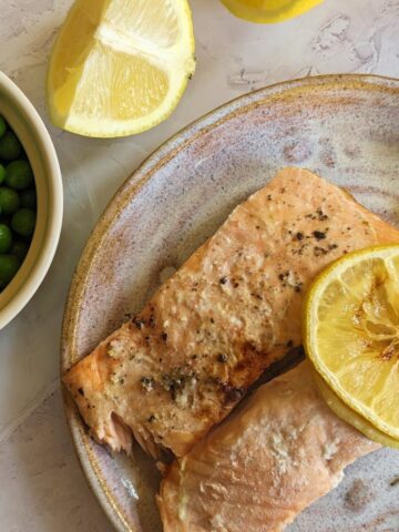 Two salmon fillets on a plate with a lemon slice on top. A bowl of peas and lemon wedges are at the side.