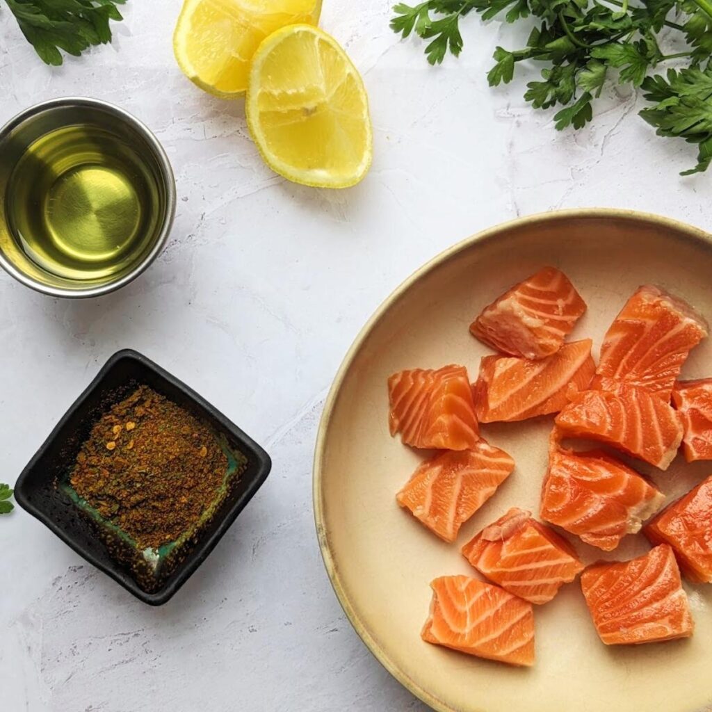 Raw salmon cut into cubes in a bowl with fresh lemon wedges, seasoning and oil at the side.