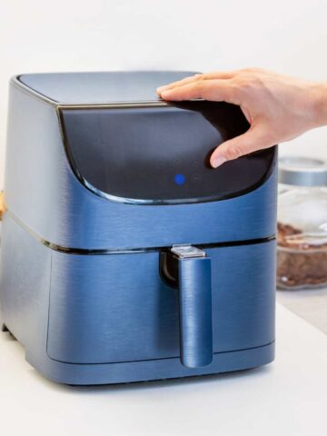 A small blue air fryer on a kitchen worktop with a hand resting on top.