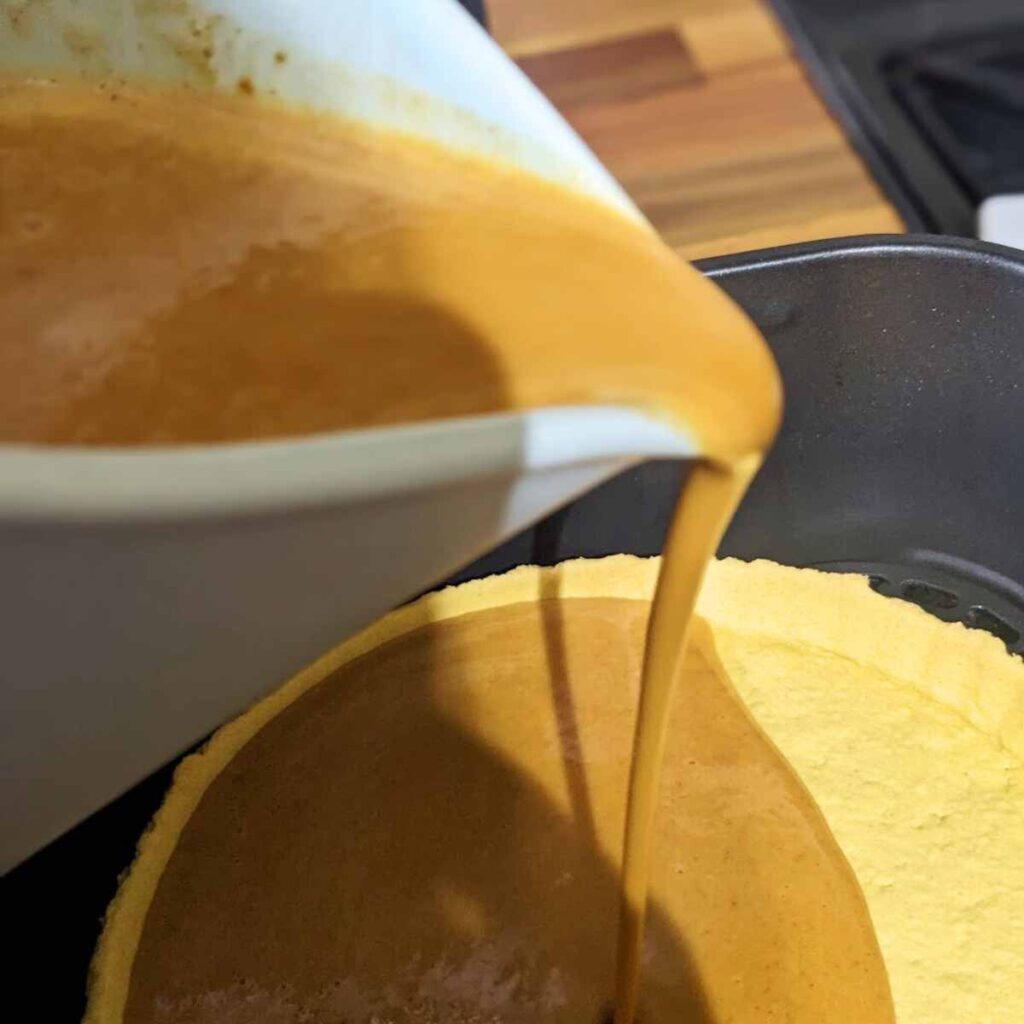 A jug of pumpkin pie filling being poured into a ready-made pie crust in an air fryer basket.