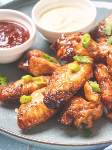 A plate of air fried chicken wings topped with slices of spring onion. Two bowls of dips sit at the side.