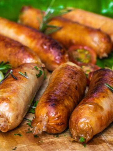 Seven cooked sausages on a wooden board with herbs scattered over.